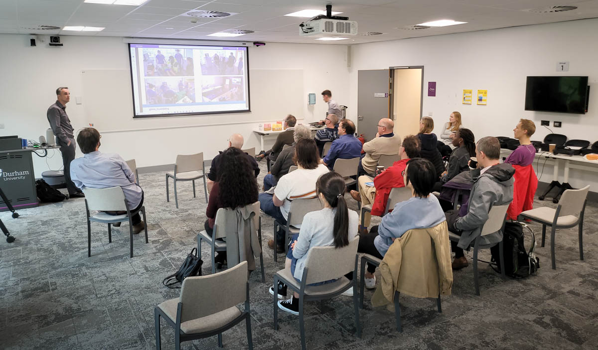 A classroom in the Teaching and Learning Centre, Durham University. A male presenter is stood beside a lectern with a projector screen. 17 people sat are sat listening, looking towards the presenter.