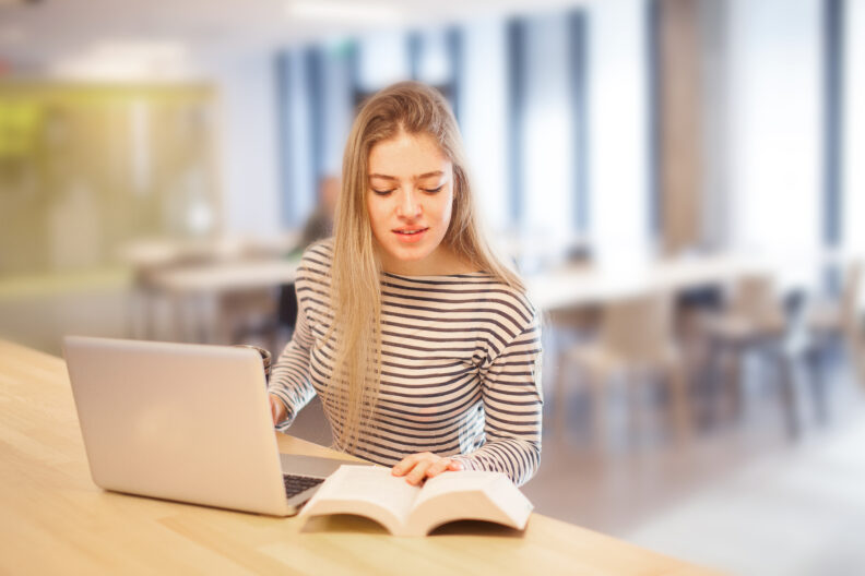 A university student with her laptop looking at a book.